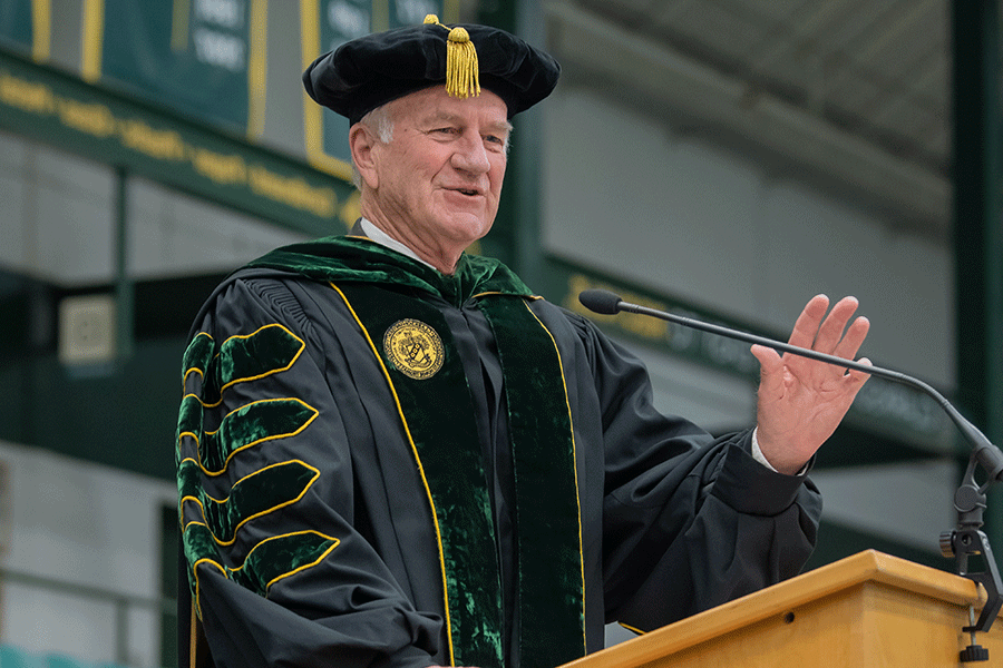 Clarkson President Emeritus Tony Collins wears regalia and speaks at a lectern during Clarkson's annual Commencement Ceremony in Cheel Arena